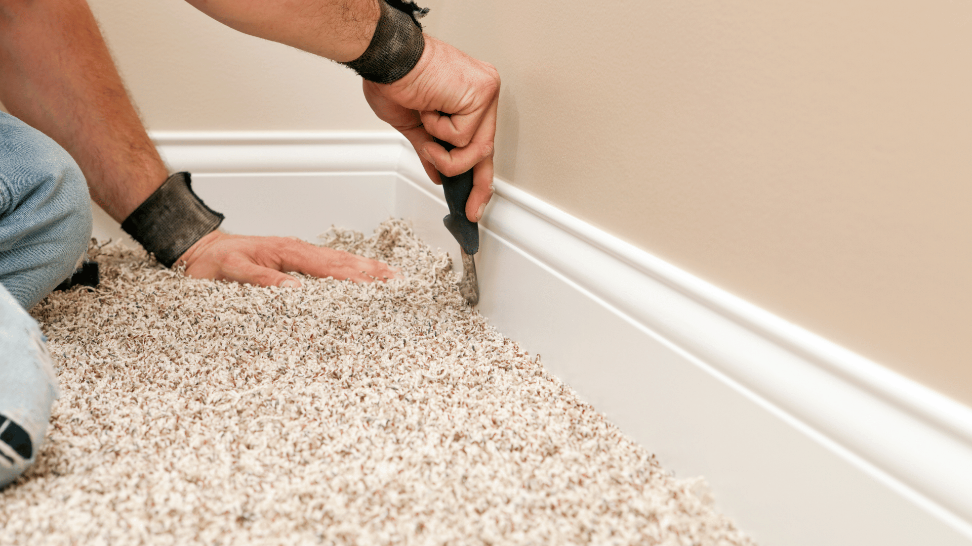 Hands using a hand tool to tuck the edge of beige plush carpet against a white baseboard