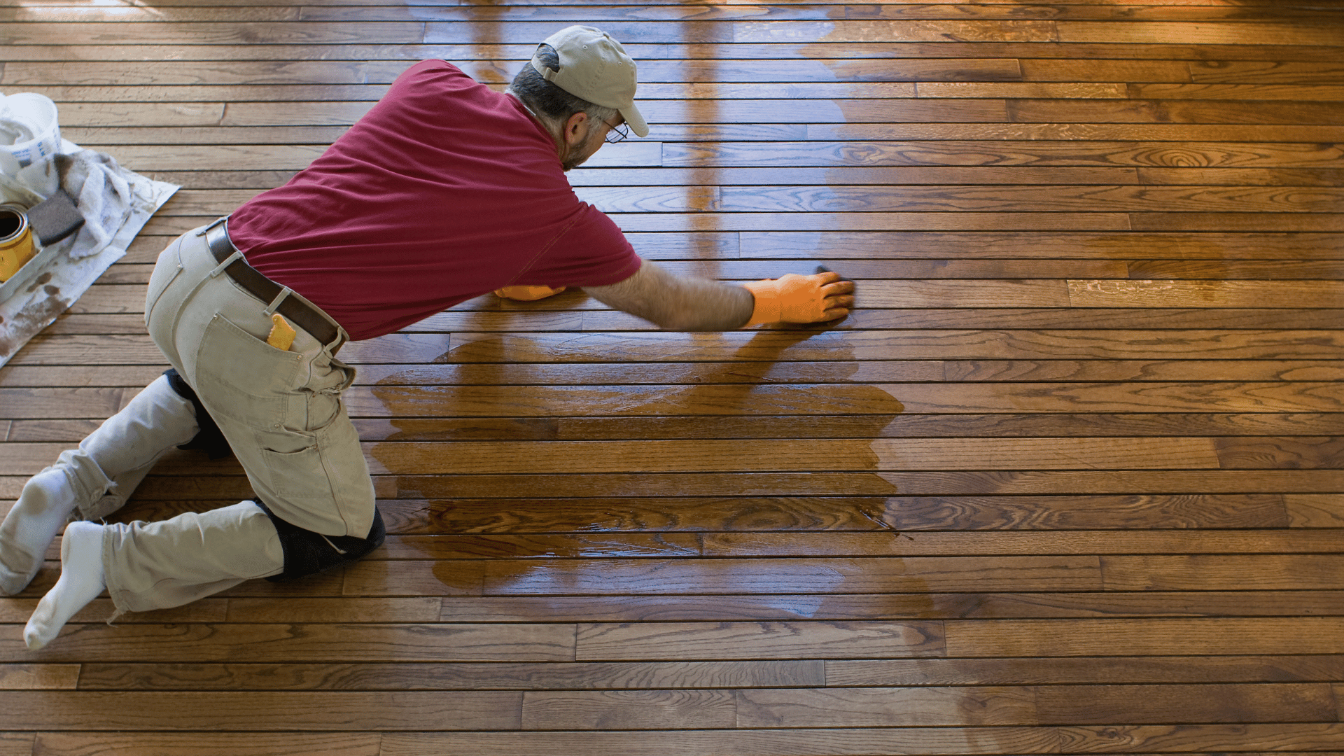 Worker in a red shirt kneeling on a hardwood floor while spreading stain with a cloth