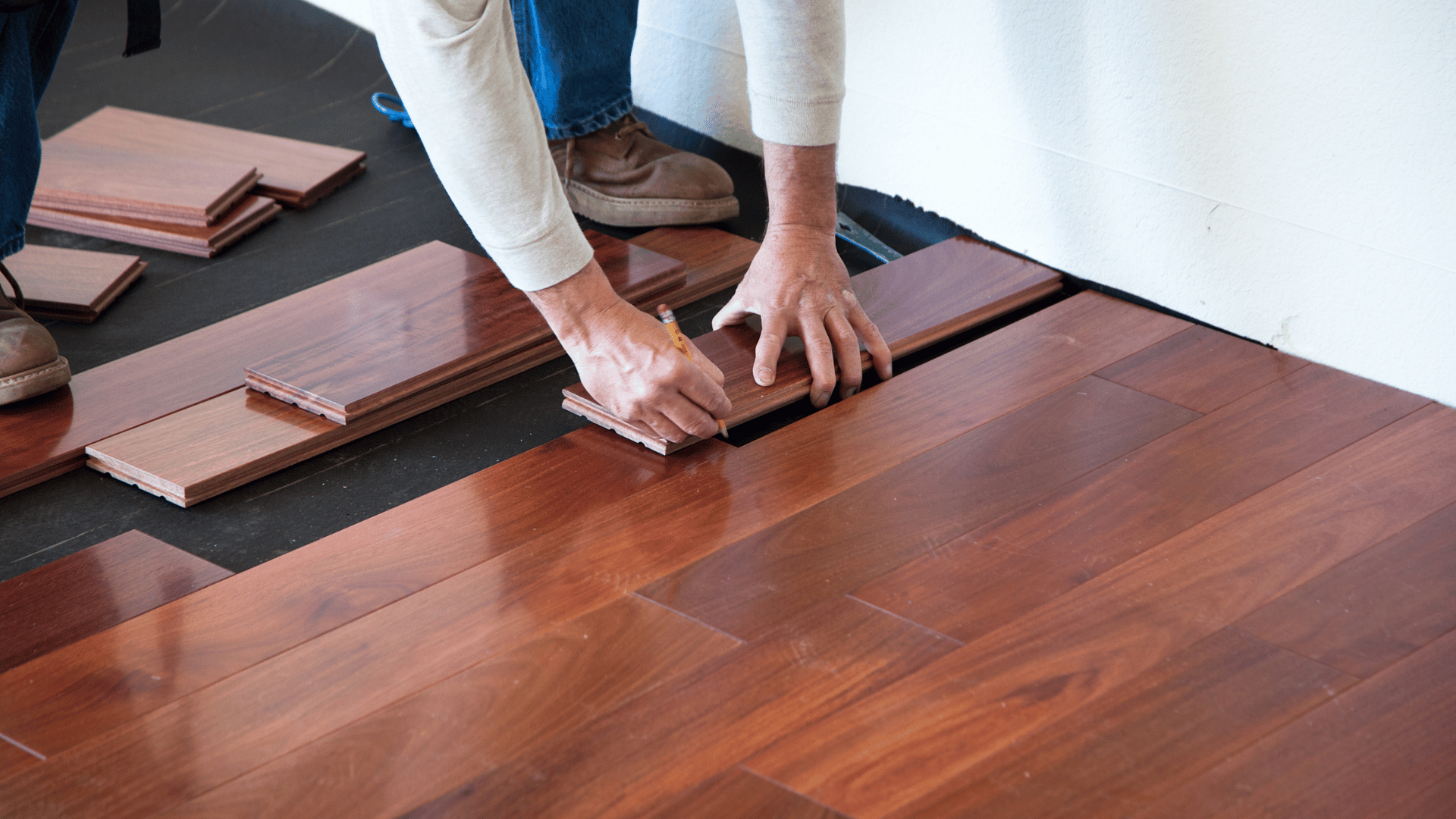 Two people's hands aligning dark reddish-brown hardwood boards as a new floor is laid