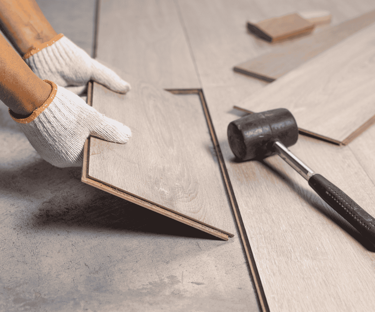 Gloved hand angling a grey wood-look plank into place next to a rubber mallet and tapping block