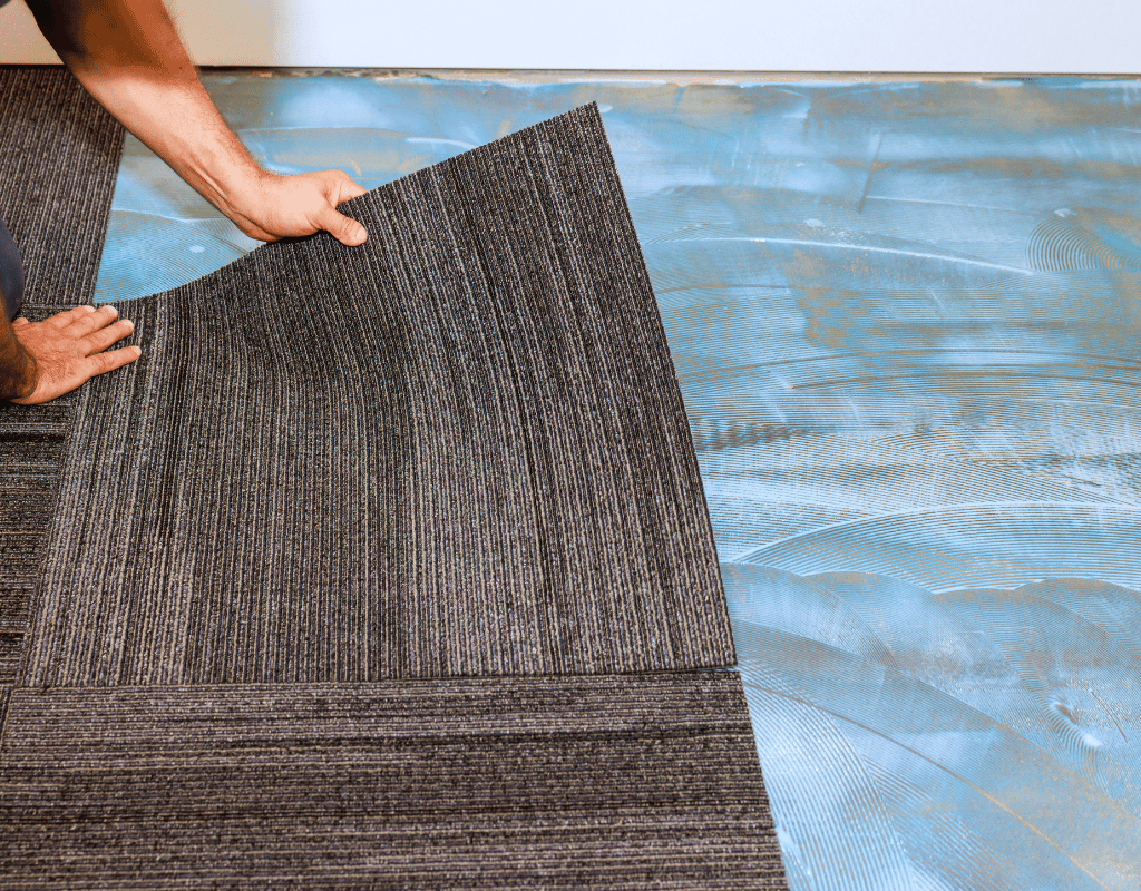 Hands positioning a dark striped carpet tile onto a subfloor coated with blue adhesive