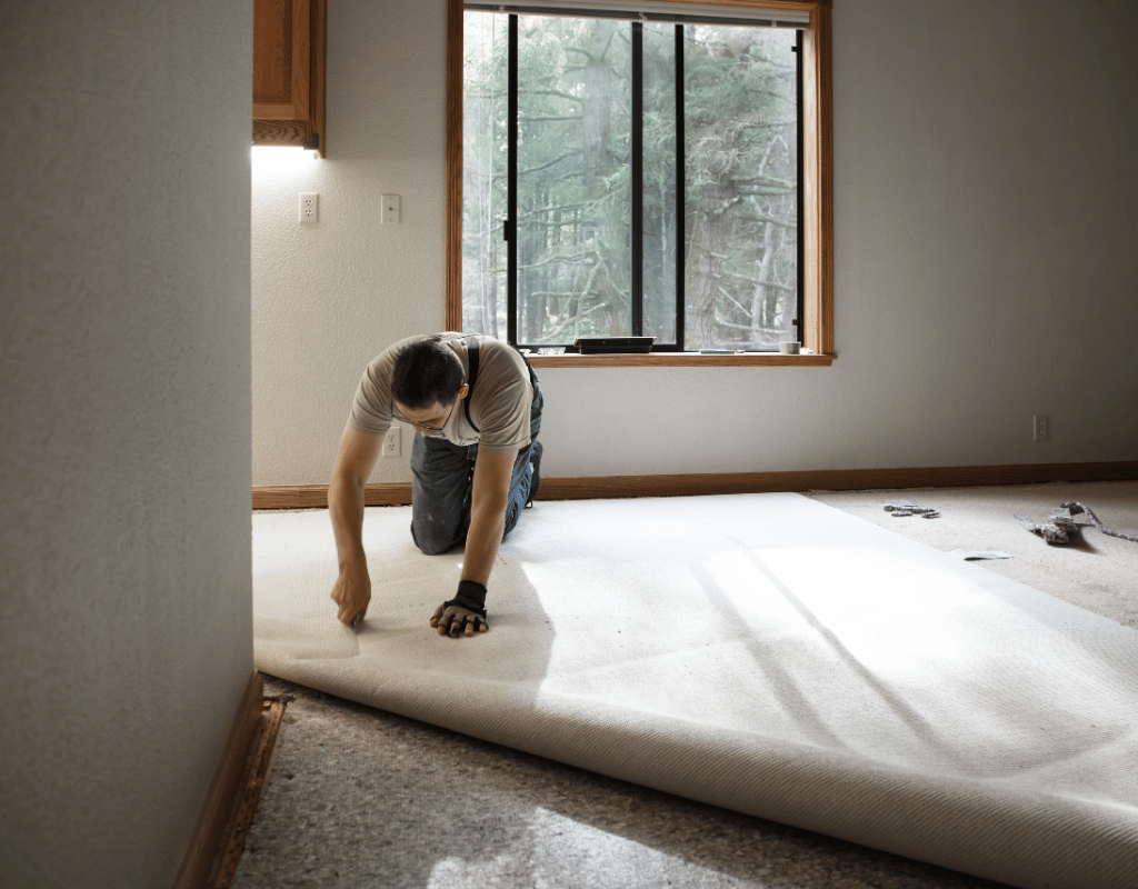 Installer kneeling on a bare floor while unrolling carpet padding across an empty room beside a window