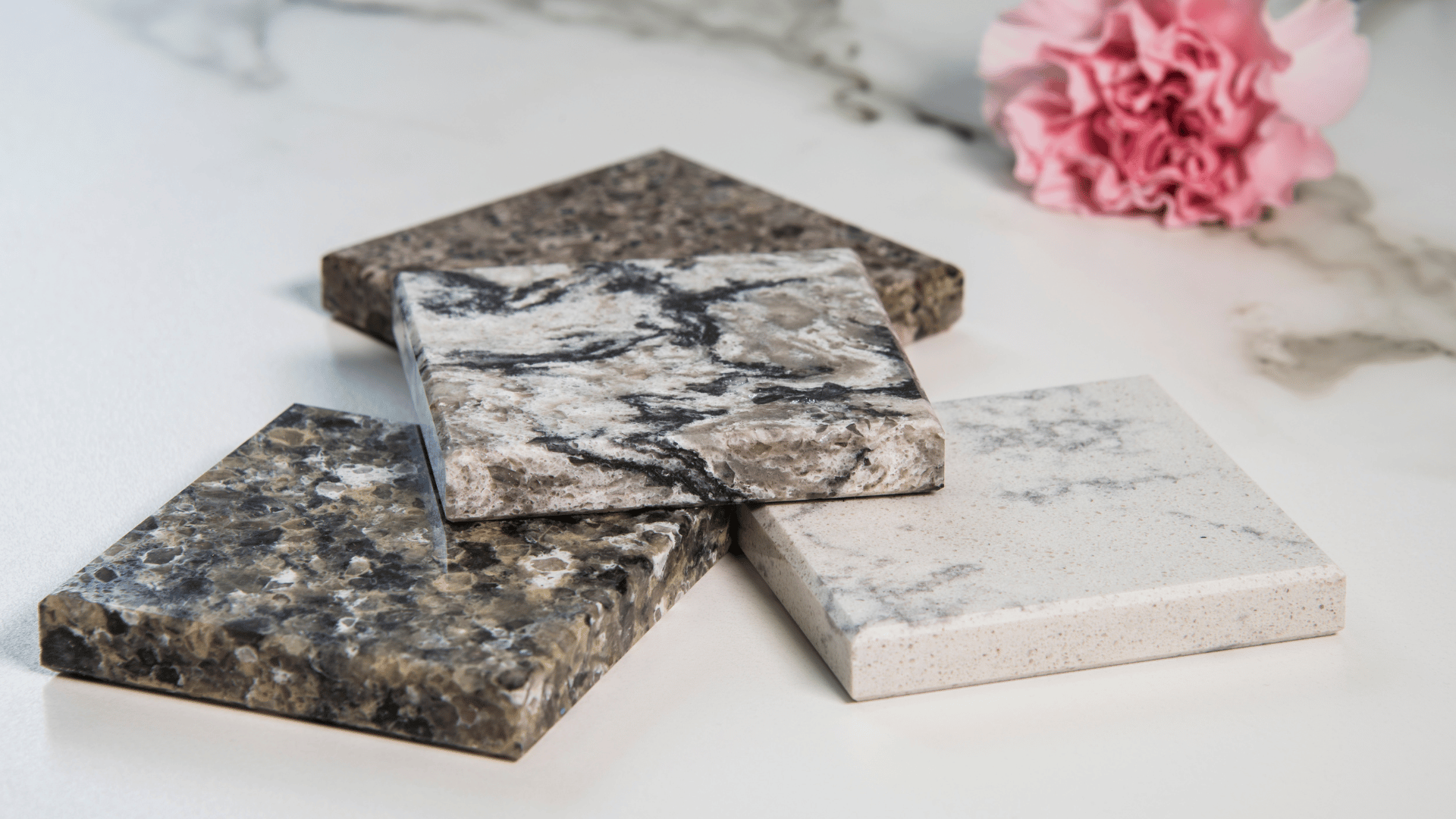Stacked squares of polished granite and marble tile resting on a white surface beside a pink carnation